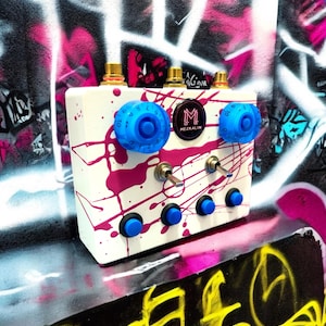 Könnte beinhalten: Ein weißes, rechteckiges Effektpedal mit einem leuchtend pinkfarbenen Farbspritzermuster. Das Gerät verfügt über zwei große blaue Drehregler, zwei Metallkippschalter, vier kleine blaue Drucktasten und ein rundes Logo-Abzeichen mit der Aufschrift MEZKALIN.