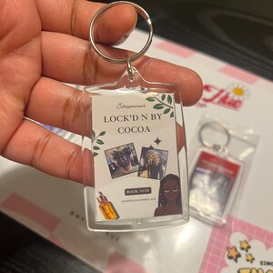 Może przedstawiać: Przezroczysty akrylowy brelok do kluczy ze srebrnym kółkiem. Brelok zawiera tekst "LOCK'D N BY COCOA" z obrazami fryzur i postacią z kreskówki. Widoczne są również słowa "BOOK NOW".