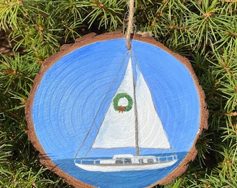 Pode incluir: Enfeite de Natal redondo em madeira com um desenho de veleiro pintado à mão. O barco tem uma vela e um casco brancos, contra um céu e mar azuis. Uma pequena grinalda verde adorna a vela. Pendurado com barbante natural.