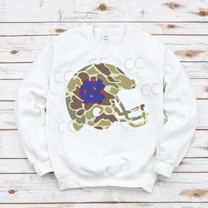 May include: White sweatshirt featuring a camouflage football helmet design with a blue and white logo. The helmet graphic is in shades of green, brown, and tan. The sweatshirt is a long-sleeved crew neck.
