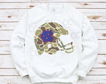 May include: White sweatshirt featuring a camouflage football helmet design with a blue and white logo. The helmet graphic is in shades of green, brown, and tan. The sweatshirt is a long-sleeved crew neck.