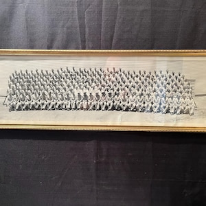 May include: A black and white photograph of a large group of people, likely military personnel, sitting in rows. They are all wearing uniforms and hats. The photo is framed in a gold frame.