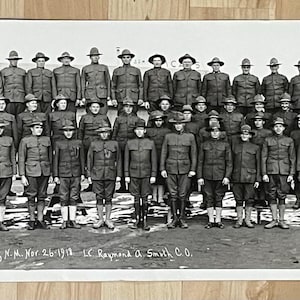 May include: A black and white photograph of a group of men in military uniform standing in a line. The men are all wearing hats and have serious expressions on their faces. The photo is likely from the early 20th century.