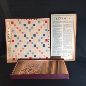 May include: A vintage Scrabble board game in its original box. The game board is brown with a cream-colored playing surface. The board has a red and blue grid pattern with a star in the center. The box is burgundy with a cream-colored label that reads "Rules for Playing Scrabble".