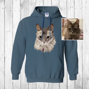 May include: A blue hooded sweatshirt with a stylized illustration of a cat on the front. The cat has gray and white fur and green eyes.