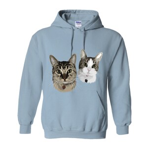 May include: A light blue hooded sweatshirt with a graphic of two cats. The cats are looking at the viewer. The cat on the left is brown and tabby, and the cat on the right is white with gray markings.