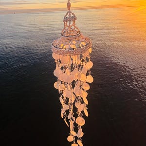 May include: A hanging seashell wind chime with a tiered design. The chime features numerous white, round shells and dangling accents. The background shows a sunset over the ocean, with the sun's glow illuminating the scene.