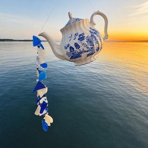 以下が含まれることがあります： 青と白の磁器のティーポットと、青と白のガラス片で作られた装飾的な風鈴。 ティーポットは、水と夕日の背景に対して空中に吊り下げられています。