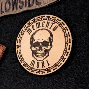 May include: A collection of leather patches and a fabric patch. The round leather patch features a skull design with the words "MEMENTO MORI" around the skull. An American flag patch and a "LOWSIDE" patch are also visible.