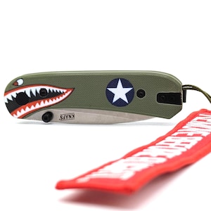 May include: A green pocket knife with a shark tooth design and a star on the handle. The knife has a silver blade and the words "KNAFS." are printed on the handle. The knife is attached to a red lanyard with white text.