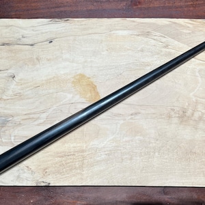May include: A long, slender, tapered black wooden wand with a smooth, polished finish. The wand is positioned diagonally across a rectangular, light-colored wooden board featuring natural grain patterns and light tan and cream color variations.