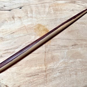 May include: A handcrafted wooden knitting needle with a pointed tip. The needle features a central stripe of dark purple wood, flanked by lighter wood tones. The needle is displayed on a light-colored wooden surface.