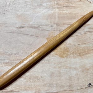 May include: A long, thin, wooden hair stick with a smooth, light brown finish. The stick is tapered at both ends, with a slightly rounded tip.