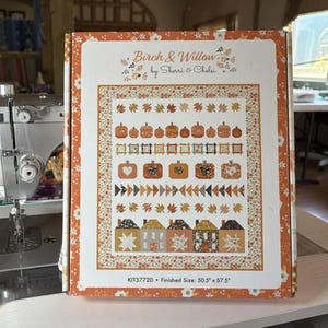 Könnte beinhalten: Eine rechteckige Schachtel für ein Birch & Willow Quilt-Set von Sherri & Chelsi. Die Schachtel hat einen weißen Hintergrund mit einem orangefarbenen Blumenrand und eine zentrale Abbildung eines Quiltmusters mit Kürbissen, Blättern und Häusern. Text: KIT37720 • Fertige Größe: 128,27 cm x 146,05 cm.