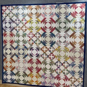 Op de afbeelding: Een handgemaakte quilt met een herhalend geometrisch patroon in verschillende kleuren. De quilt heeft vierkanten en ruitvormen in tinten rood, blauw, groen, geel en paars, tegen een witte achtergrond. De quilt is afgezet met een donkerblauwe stof.