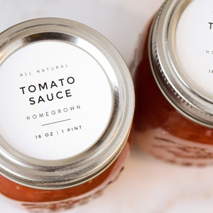 May include: Two glass jars with silver lids filled with tomato sauce. The jars have white labels with black text that reads "All Natural Tomato Sauce Homegrown 16 oz | 1 Pint".