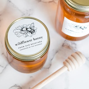May include: Two glass jars of honey with gold lids and white labels. The labels have a black and white illustration of a bee and a flower. The text on the label reads "wildflower honey from our backyard harvested by the Jacksons". A wooden honey dipper is in the foreground.