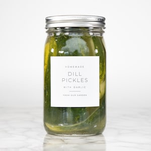 May include: A glass jar filled with green dill pickles and a white label that reads "HOMEMADE DILL PICKLES WITH GARLIC FROM OUR GARDEN".