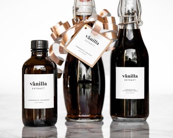 May include: Three glass bottles of vanilla extract with labels that say "vanilla extract" in black text on a white background. The bottles are on a white marble surface. The bottle in the center is tied with a gold ribbon.