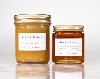 May include: Two glass jars of clover honey, one larger than the other. Both jars have gold lids and white labels with the text "clover honey" and "from our backyard bees". The smaller jar is filled with a darker honey, while the larger jar is filled with a lighter honey.