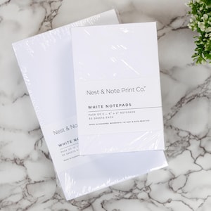 May include: Two packs of white notepads, each with 50 sheets. The notepads are wrapped in plastic and have the text "Nest & Note Print Co." and "White Notepads" printed on the front.