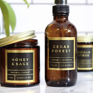 May include: Two amber glass bottles with gold labels. The bottle on the left is a 4 ounce beeswax candle labelled "Honey & Sage". The bottle on the right is a 4 fluid ounce room spray labelled "Cedar Forest" and "Parfum d'Intérieur". Both bottles have black text on gold labels.