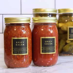 May include: Two glass jars of red salsa with gold lids and black labels that read "Spicy Salsa with Serrano Chiles" and "All Natural".  A glass jar of green pickles with a gold lid and a black label that reads "Spicy Dills". A small glass bottle with a black lid and a black label that reads "Vanilla Extract".