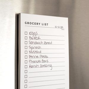 May include: A white grocery list notepad with a black border is attached to a stainless steel surface. The list includes items like eggs, butter, and spinach. The date "10-10-25" is written at the top.