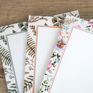 May include: A stack of floral-themed notepads with botanical designs. The notepads feature various floral and leaf patterns in shades of green, brown, pink, and white. One notepad has the word "notes" written in a script font.