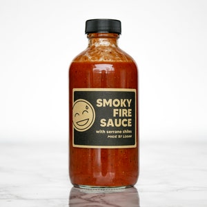 May include: A glass bottle of "Smoky Fire Sauce" with a black cap. The label features a smiling face graphic and text that reads "Smoky Fire Sauce with serrano chiles MADE BY LOGAN". The sauce is a reddish-brown colour.