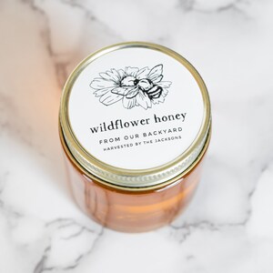 May include: A glass jar of wildflower honey with a gold lid and a white label that reads "wildflower honey from our backyard harvested by the Jacksons". The label also features a black and white illustration of a bee on a flower.