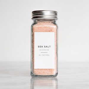 May include: A glass jar with a silver lid filled with pink sea salt. The jar has a white label that reads "SEA SALT SEASONING ALL NATURAL".