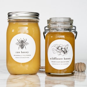 May include: Two glass jars of honey with white labels. The jar on the left is labeled "raw honey" with a bee illustration. The jar on the right is labeled "wildflower honey" with a bee and flower illustration. Both labels state "minimally filtered" and "from our backyard".