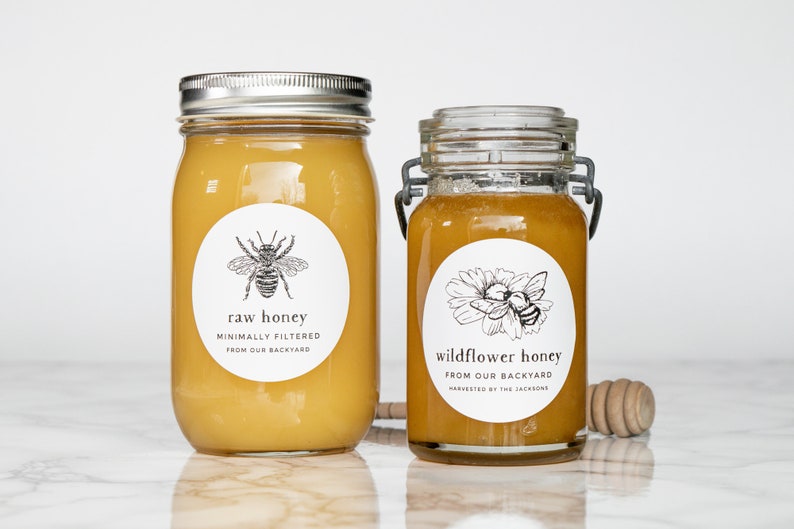 May include: Two glass jars of honey with white labels. The jar on the left is labeled "raw honey" with a bee illustration. The jar on the right is labeled "wildflower honey" with a bee and flower illustration. Both labels state "minimally filtered" and "from our backyard".