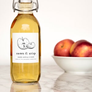 Może przedstawiać: Przezroczysta szklana butelka z etykietą, na kt&oacute;rej widnieje napis "sweet & crisp hard apple cider made at the orchard homestead". Etykieta zawiera czarno-biały rysunek jabłka. W tle znajdują się trzy czerwone jabłka w białej misce.