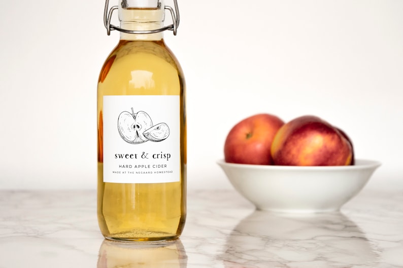Może przedstawiać: Przezroczysta szklana butelka z etykietą, na kt&oacute;rej widnieje napis "sweet & crisp hard apple cider made at the orchard homestead". Etykieta zawiera czarno-biały rysunek jabłka. W tle znajdują się trzy czerwone jabłka w białej misce.