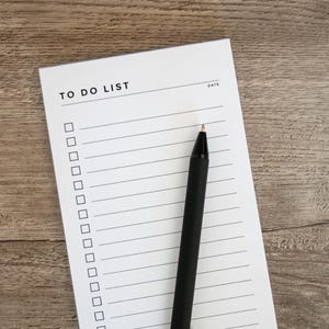 May include: A white notepad with the words "TO DO LIST" printed at the top, featuring a series of checkboxes and lined spaces. A black pen rests diagonally across the notepad. The background is a wooden surface.