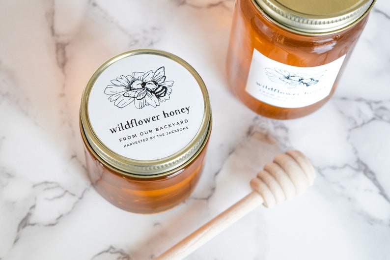 May include: Two glass jars of honey with gold lids and white labels. The labels have a black and white illustration of a bee and a flower. The text on the label reads "wildflower honey from our backyard harvested by the Jacksons". A wooden honey dipper is in the foreground.