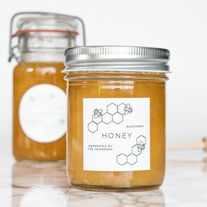 May include: Two glass jars of honey with white labels. The label on the front jar reads "Backyard Honey, Harvested by The Johnsons". The label features a black and white illustration of a honeycomb with bees.
