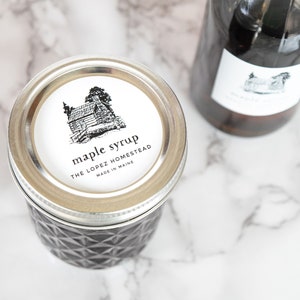 May include: A glass jar with a silver lid filled with maple syrup. The jar has a black and white label that reads "maple syrup" and "The Lopez Homestead, Made in Maine".