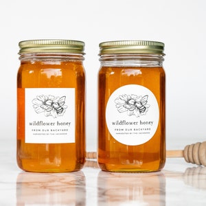 May include: Two glass jars filled with honey, labeled "wildflower honey from our backyard, harvested by the Jackson's". The jars have gold lids and are sitting on a white marble surface. A wooden honey dipper is in the foreground.