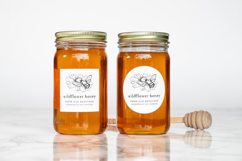 May include: Two glass jars filled with honey, labeled "wildflower honey from our backyard, harvested by the Jackson's". The jars have gold lids and are sitting on a white marble surface. A wooden honey dipper is in the foreground.