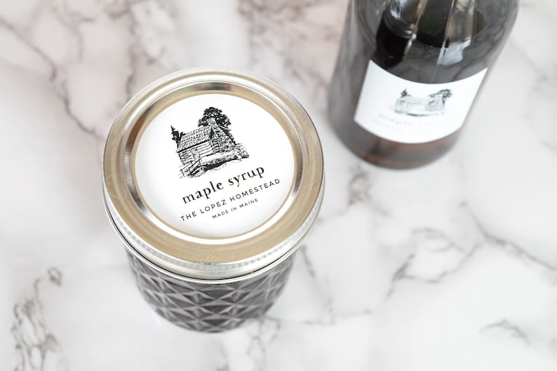 May include: A glass jar with a silver lid filled with maple syrup. The label on the jar reads "maple syrup, The Lopez Homestead, Made in Maine". The label also features a black and white illustration of a cabin.