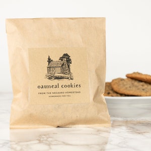 Może przedstawiać: Brązowa papierowa torba z etykietą z napisem "oatmeal cookies from the Negaard Homestead homemade for you". Etykieta zawiera czarno-białą ilustrację chaty w lesie.