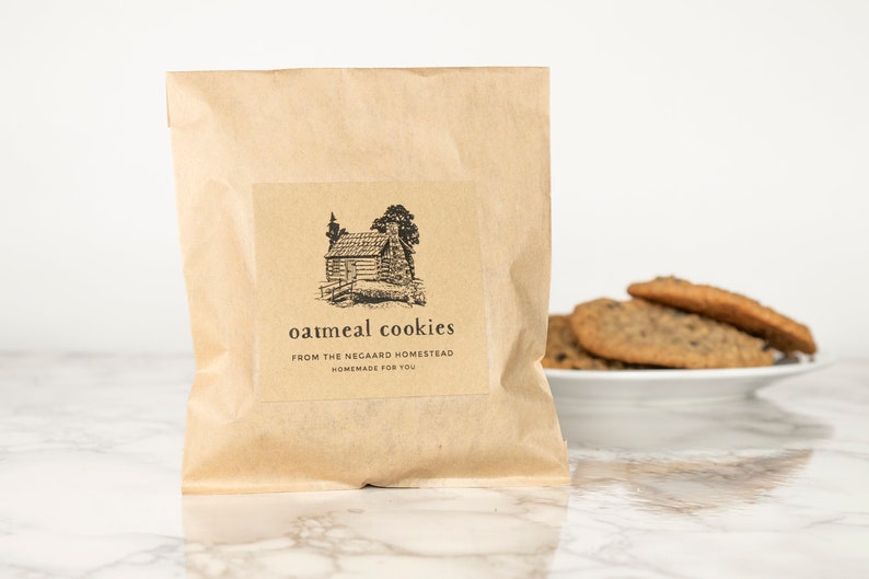 Może przedstawiać: Brązowa papierowa torba z etykietą z napisem "oatmeal cookies from the Negaard Homestead homemade for you". Etykieta zawiera czarno-białą ilustrację chaty w lesie.
