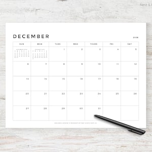 May include: A white paper calendar for December 2026 with a minimalist design. The calendar grid is black, with the month's name at the top. A black pen rests on the bottom right corner. The calendar is on a light wood surface.