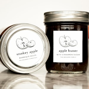 Może przedstawiać: Dwa szklane słoiki z białymi etykietami i czarnymi ilustracjami jabłek. Etykiety głosiły "smokey apple barbeque sauce the negaard homestead" i "apple butter with cinnamon sugar the negaard family".