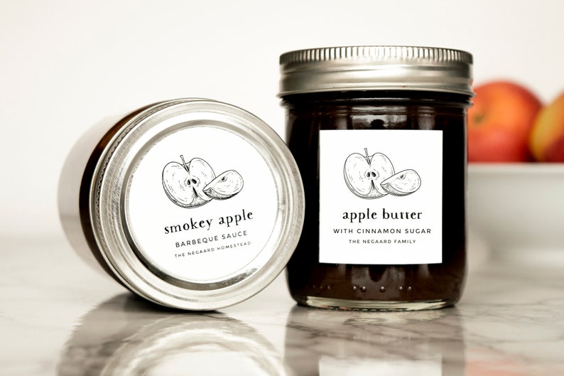 Może przedstawiać: Dwa szklane słoiki z białymi etykietami i czarnymi ilustracjami jabłek. Etykiety głosiły "smokey apple barbeque sauce the negaard homestead" i "apple butter with cinnamon sugar the negaard family".