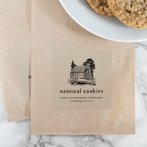 May include: A brown paper bag with a sticker that says "oatmeal cookies from the Negaard Homestead homemade for you". The sticker features a black and white illustration of a cabin in the woods.