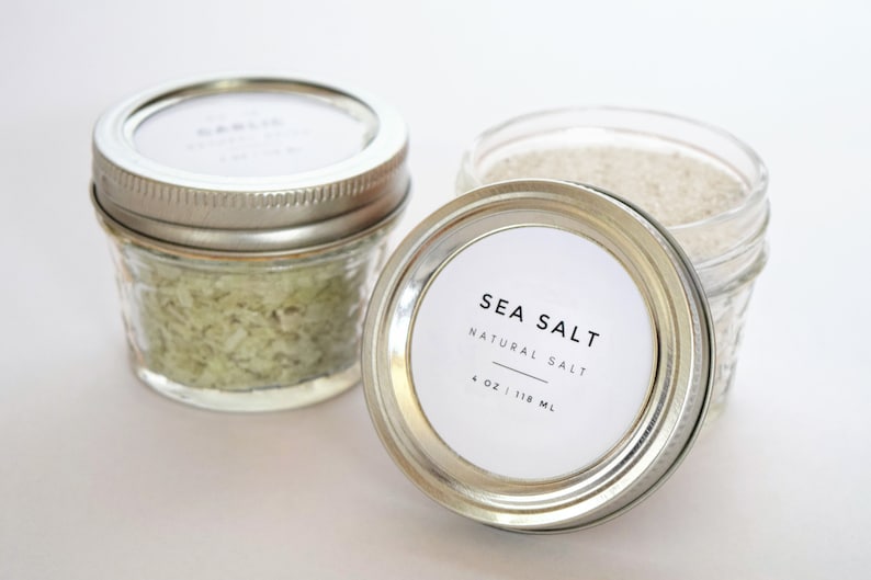 May include: Two glass jars with silver lids filled with sea salt. One jar has a white label that reads "SEA SALT NATURAL SALT 4 OZ | 118 ML".